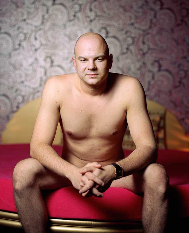 Mann namens Ralf sitzt nackt auf einem roten Bett mit verschränkten Händen und trägt eine Armbanduhr